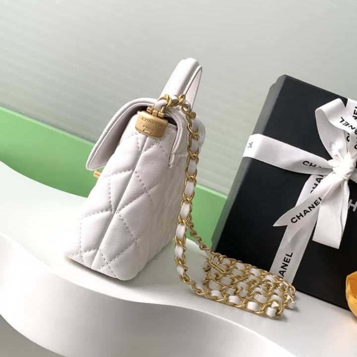 White quilted handbag with gold chain and black Chanel box with white ribbon on a white surface.